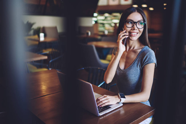 Cheerful woman enjoying telephone conversation during time for remote work on netbook, successful female freelancer calling to friend for discussing ideas about project with laptop application - Photo, Image