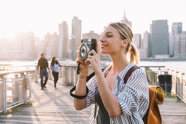Carefree female tourist with stylish rucksack standing at urban setting with Manhattan district on background and focusing lens of instant camera for clicking pictures, concept of recreating - Foto, imagen
