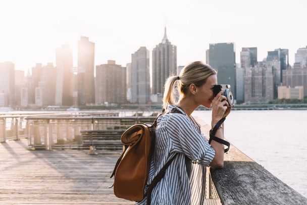 Side view of skilled hipster girl with instant camera clicking picture of city landscape standing at urban setting with Manhattan view, positive female tourist with backpack enjoying getaway - Foto, imagen
