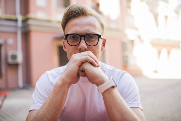 Close up portrait of handsome hipster guy in optical spectacles for vision correction looking at camera during leisure time outdoors, serious man in classic glasses sitting at urban setting - Zdjęcie, obraz