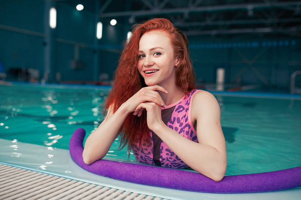 Beautiful young red-haired girl in a fashionable swimsuit in the style of the 80s stands with the noodles in her hands in the indoor swimming pool - Foto, Bild