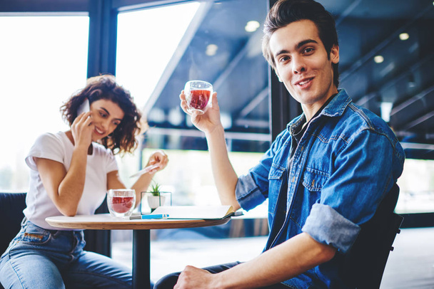 Portrait of handsome young man dressed in casual wear holding glass of juice and looking at camera while cheerful young woman calling on mobile phone sitting at table in coffee shop - Foto, immagini