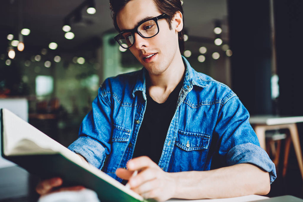 Concentrated hipster guy in optical spectacles reading knowledge literature in coffee shop for making research of information for course work, handsome man holding textbook for education in hands - Fotoğraf, Görsel
