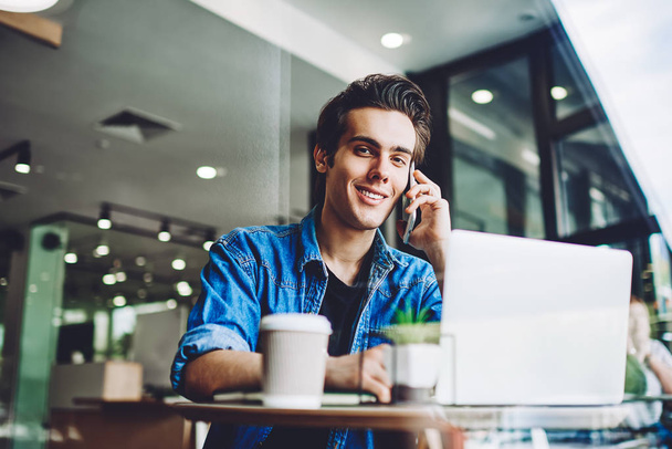 Positive skilled hipster guy communicated with colleague via application on smartphone gadget while sitting at table with laptop device, smiling man enjoying of good wireless internet for calling - Foto, imagen