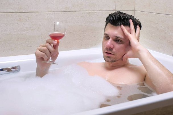 Frustrated depressed man is lying in bath and drinking wine. - Фото, зображення