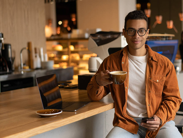 Portrait of stylish African American man freelancer holding cup of coffee and using smartphone and mobile application, looking at camera and smiling, sitting in modern cafe. Coffee break concept - Foto, afbeelding