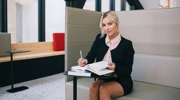 Portrait of beautiful manager holding pen in hand and looking at camera while sitting with books in workshop with modern interior, successful executive female entrepreneur spending time for knowledge - Photo, image