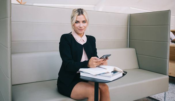 Portrait of successful female owner of small business looking at camera while waiting for meeting with partner in workspace, positive woman financial expert holding smartphone device sitting at table - Fotó, kép