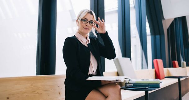 Portrait of blonde woman in fashionable optical spectacles with literature in hand sitting in modern workspace, attractive female boss dressed in formal wear holding book with management information - Photo, image