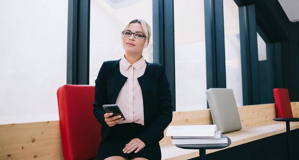 Portrait of confident female owner of business dressed in formal elegant suit sitting in office, woman in formal in stylish spectacles holding modern cellular phone and looking at camera indoors - Foto, Bild