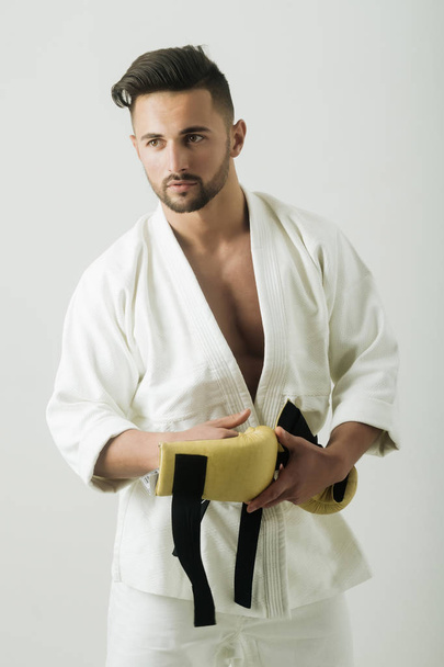 Sexy athlete, karatek with black belt. Strength and attractiv, perfect guy on white background. Kimono on the bare chest. Bearded stylish athlete before the fight. - Photo, Image