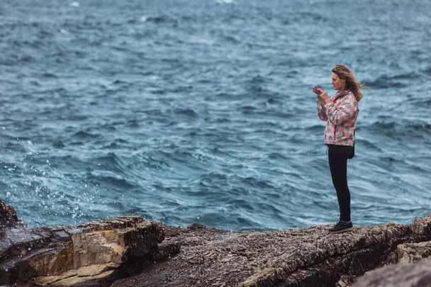 mladá žena v kabátě na pobřeží a dívá se na bouřující moře - Fotografie, Obrázek
