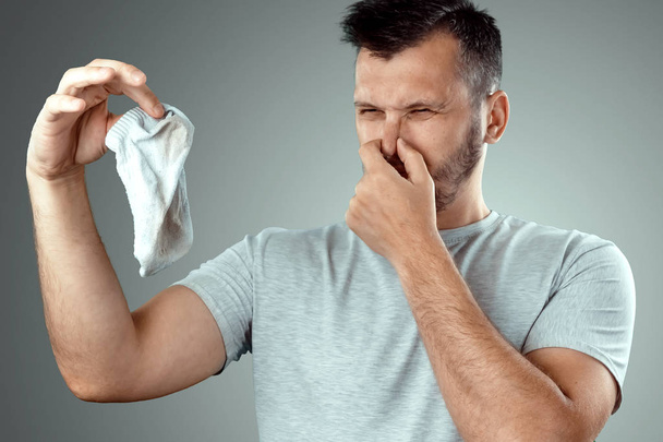 A young man holds his smelly socks and covers his nose with his hand. Nasty smell. Stink legs, fungus on the legs - Photo, image