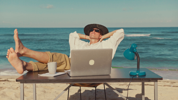 Ένας νεαρός άντρας κάθεται στην παραλία με φορητό υπολογιστή, ξεκουράζεται βάζοντας τα πόδια του στο τραπέζι - Πλάνα, βίντεο