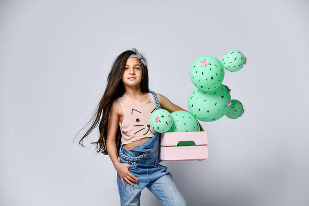Young brunette girl hold cactus balloon in pastel pink wooden box. Flowers are made of balloons with pink flowers and painted spines. Creative idea minimal concept on gray background - Valokuva, kuva