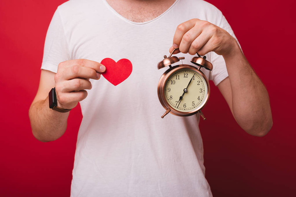 Beyaz gömlekli bir adam's tors önünde küçük bir çalar saat ve kırmızı bir kağıt kalp tutan iki el . Zaman yönetimine bayılırım. Aşk zamanı. - Fotoğraf, Görsel