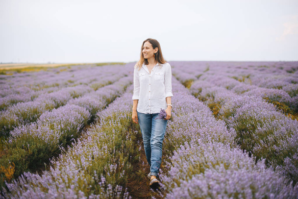 Young cheerful woman is walking on lavender field smiling and looking far away.  - Valokuva, kuva