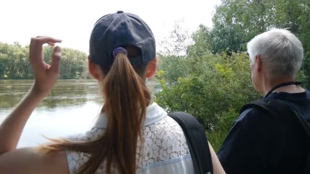 Чоловік і дівчинка ходять разом. Туристи з рюкзаками. Вони зупиняються на березі річки і дивляться на неї. Дівчина торкається і виправляє довге волосся
. - Кадри, відео