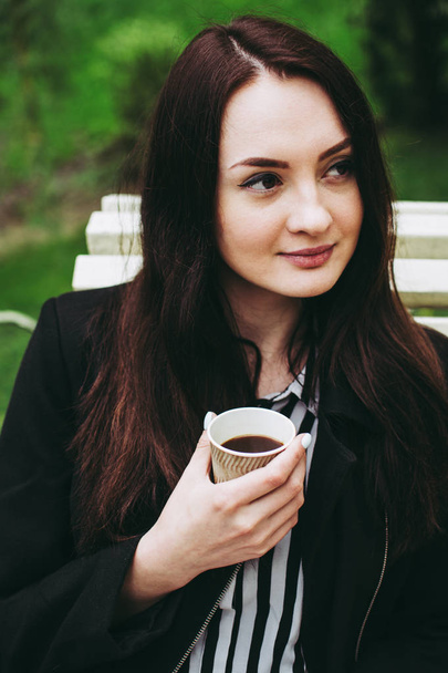 Cute brunetka uśmiechnięta trzyma kubek papieru czarnej kawy i patrząc w odległości. Młoda kobieta ubrana w czarną warstwę i pasiasty czarno-białą koszulę. - Zdjęcie, obraz