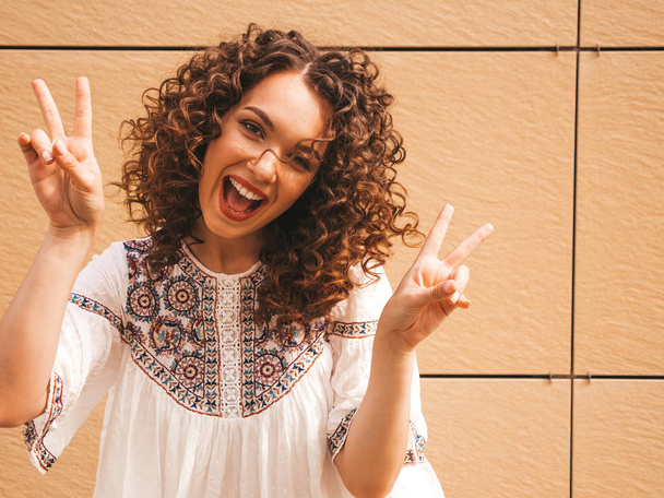 Beautiful smiling model with afro curls hairstyle dressed in summer hipster white dress.Sexy carefree girl posing in the street near yellow wall.Funny and positive woman having fun.Shows peace sign - Photo, Image