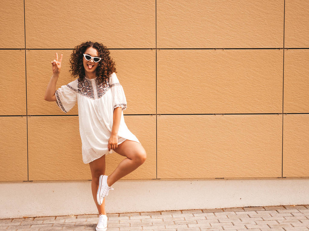 Beautiful smiling model with afro curls hairstyle dressed in summer hipster white dress.Sexy carefree girl posing in street near yellow wall in sunglasses.Positive woman having fun.Full length - Foto, Imagen