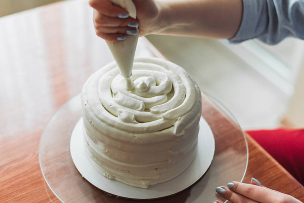Una chica encantadora haciendo un pastel en una panadería. La chica suaviza la crema del pastel. Pastel blanco en una mesa de madera
. - Foto, Imagen