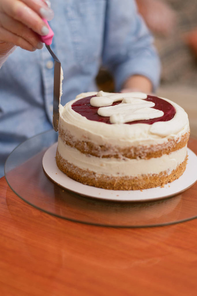 Милая девушка делает торт в пекарне. Пекарь сжимает крем на пустом красном торте. Белый торт на деревянном столе
. - Фото, изображение