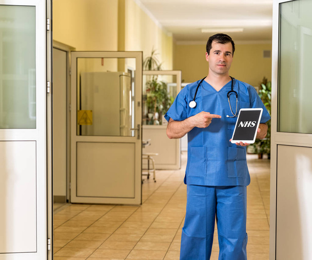 Mužský doktor středního věku v modrých šrvkách drží a míří k tabletu s textem NHS - Fotografie, Obrázek