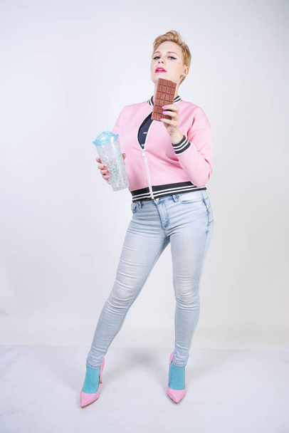 pretty plus size adult girl with short hair love chocolate. cheerful young woman enjoys sweet food. cute curvy lady in pink sport jacket holds chocolate bar in her hands on white studio background. - Fotó, kép