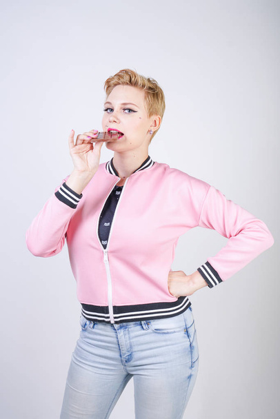 pretty plus size adult girl with short hair love chocolate. cheerful young woman enjoys sweet food. cute curvy lady in pink sport jacket holds chocolate bar in her hands on white studio background. - Photo, Image