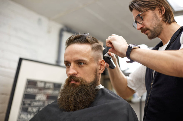 Um brutamontes na Barbearia moderna. Cabeleireiro faz penteado um homem com uma longa barba. Cabeleireiro mestre faz penteado com cortador de cabelo
 - Foto, Imagem