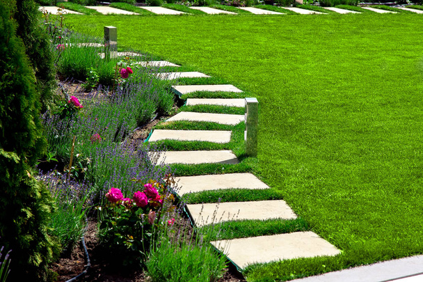 hátsó udvarban egy márvány kő sétány négyzet alakú csempe közelében egy zöld rét egy virágágyóval, kerti részletek növényekkel egy napsütéses nyári napon. - Fotó, kép