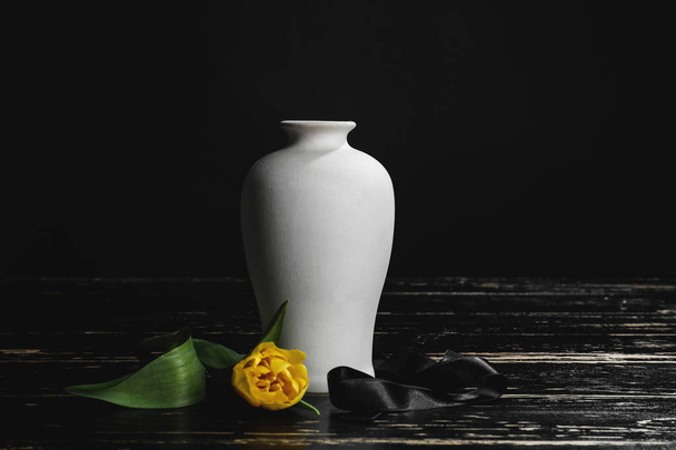 Mortuary urn with ribbon and flower on table against dark background - Photo, Image