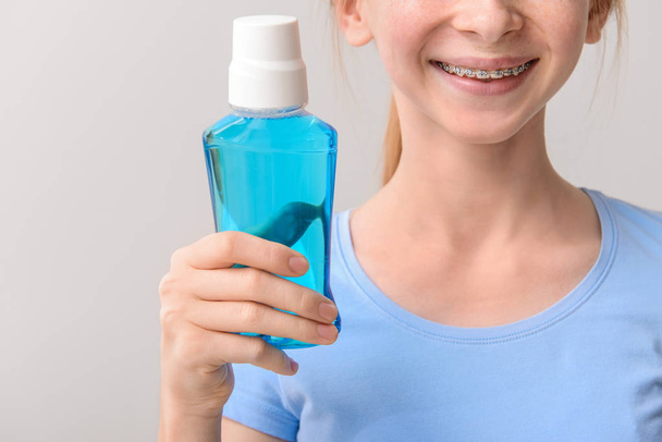 Улыбающаяся девочка-подросток с зубными скобками, держащая бутылку жидкости для полоскания рта на сером фоне
 - Фото, изображение