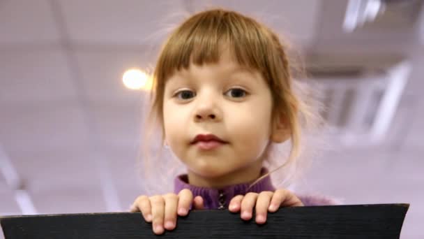 klein meisje kijkt uit achter een stoel en grimaces - Video