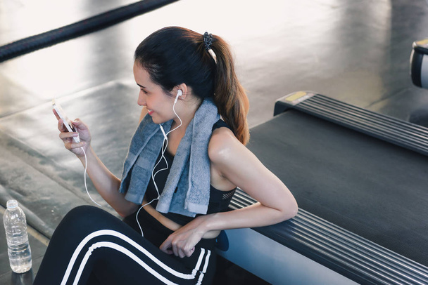 Женщина пользуется смартфоном после тренировки в фитнес-клубе, портрет женщины, слушающей музыку в наушниках во время занятий в тренажерном зале, концепция занятий спортом и досугом
. - Фото, изображение