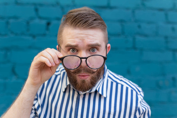 Retrato de close-up de um jovem curioso surpreso em óculos com bigode e barba posando em um fundo de parede de tijolo borrada azul. Conceito de surpresa e informação chocante
. - Foto, Imagem