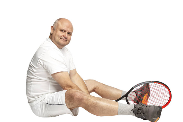 A middle aged man sits on the floor with a tennis racket in his hands. Isolated on a white background. - Foto, afbeelding