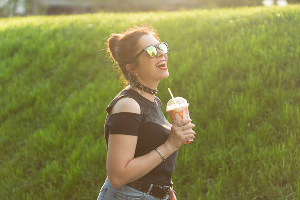 Positive cheerful young woman in stylish punk clothes and glasses holding a milkshake in her hands during a walk in the park on a warm summer evening. The concept of relaxation and fun in the city. - Photo, Image