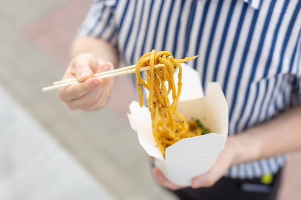 Hands of an unidentified young man in a striped shirt holding a box of udon noodles in the street in the park. Concept of Asian cuisine. Takeaway food. - Photo, Image