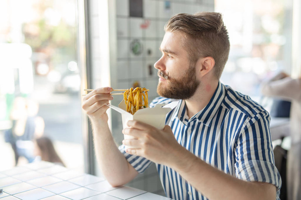 Vista laterale di un ragazzo piuttosto giovane hipster mangiare tagliatelle udon utilizzando bacchette di legno seduto in un caffè e guardando nella finestra. Concetto cucina asiatica
. - Foto, immagini