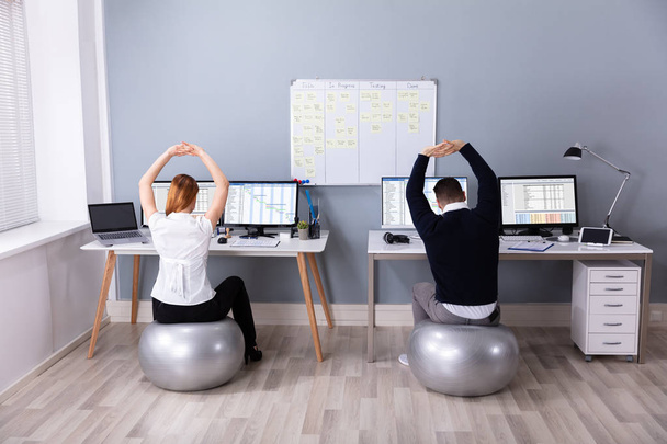 Rear View Of Businesspeople Sitting On Fitness Ball With Arm Raised At Workplace - Photo, image