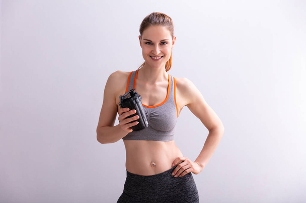 Portrait Of Smiling Fitness Woman Holding Protein Shaker Bottle Against Gray Background - Fotografie, Obrázek