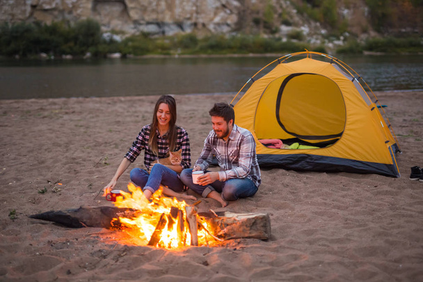 people, summer tourism and nature concept - couple near camp fire warming up seen from the tent - Photo, Image