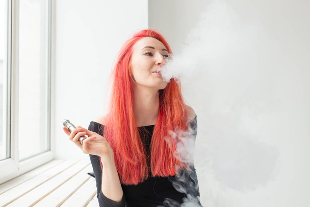 Jugend- und Suchtkonzept - junge rothaarige Frau raucht Dampf am Fenster - Foto, Bild