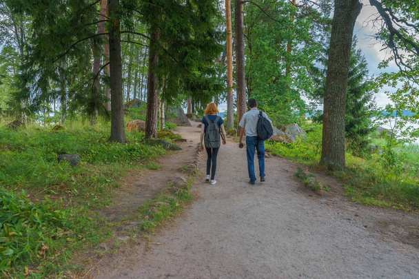 Dark-haired middle-aged man and red-haired young lady walk along forest road. Tourists on the beautiful landscape background. Monrepos Park, Vyborg, Russia. Travel concept - Foto, Imagen