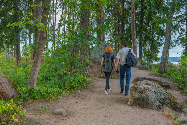 Dark-haired middle-aged man and red-haired young lady walk along forest road. Tourists on the beautiful landscape background. Monrepos Park, Vyborg, Russia. Travel concept - Фото, зображення