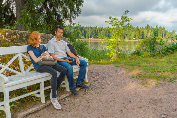 Handsome middle-aged man and young pretty lady sitting on bench and using smartphones in summer evening. Tourists on the beautiful landscape background. Monrepos Park, Vyborg, Russia. Travel concept - Photo, Image