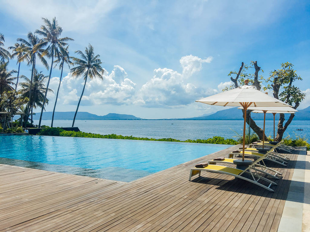 Piękny tropikalny basen w hotelu lub ośrodku z parasolem, kokosy drzewa leżaki, palmy z widokiem na basen bez krawędzi, ocean i górskie tło - Zdjęcie, obraz