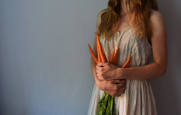 Mujer sosteniendo un montón de zanahorias orgánicas frescas. Chica de pelo largo sonriente en el vestido blanco largo. Las verduras en las manos
. - Foto, Imagen